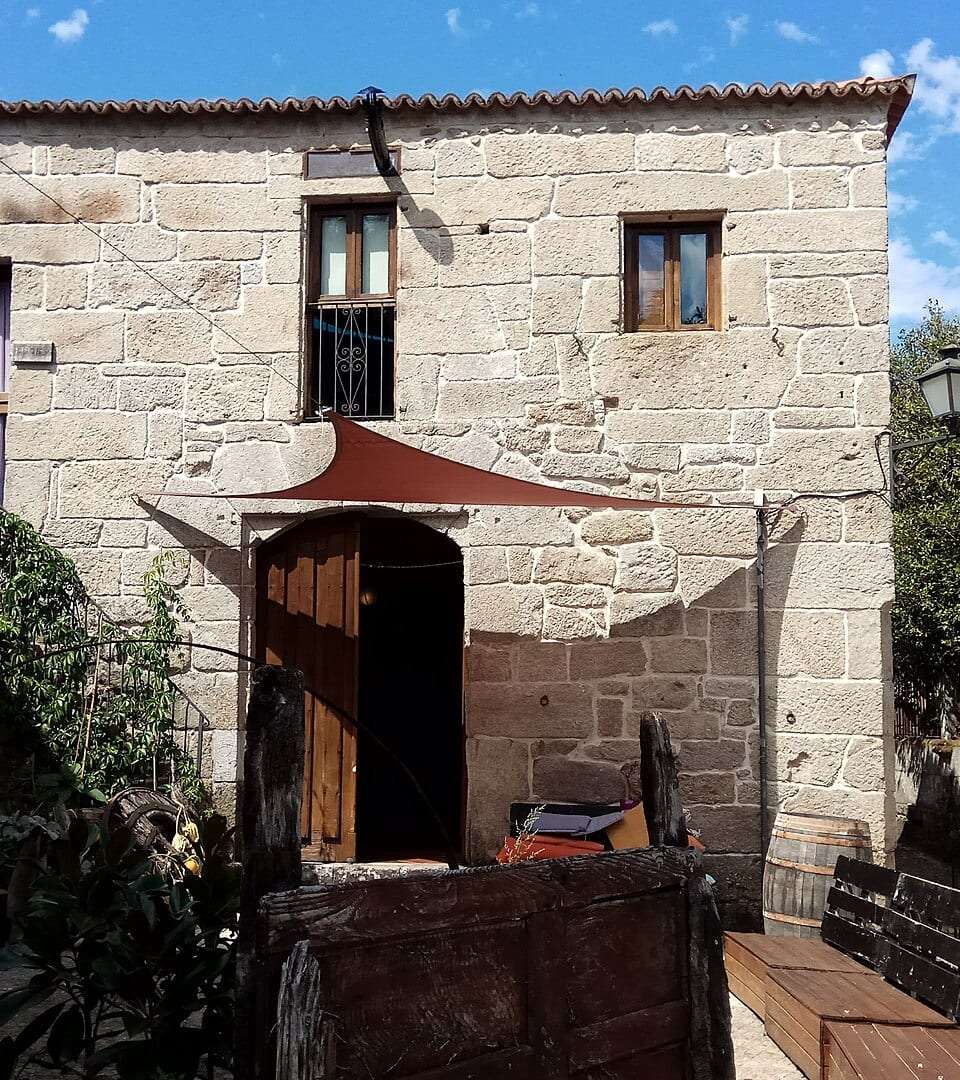 Fachada de piedra de una casa rural con toldo y banco de madera en la entrada, en Pazos de Arenteiro